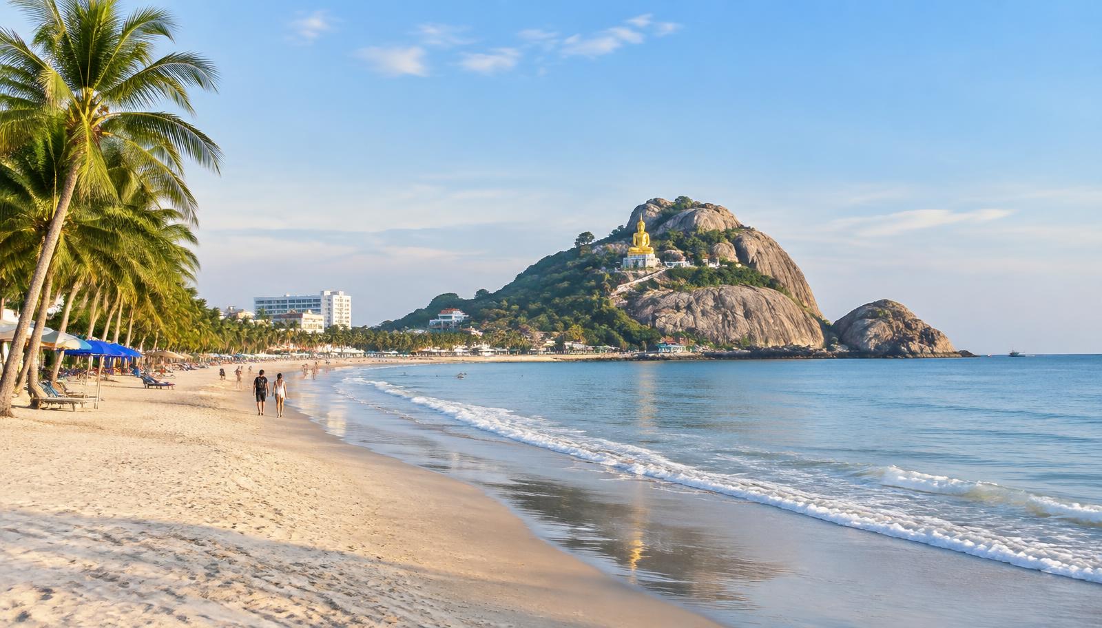 Hua Hin beach with palm trees and Khao Takiab hill with its golden Buddha statue in soft morning light